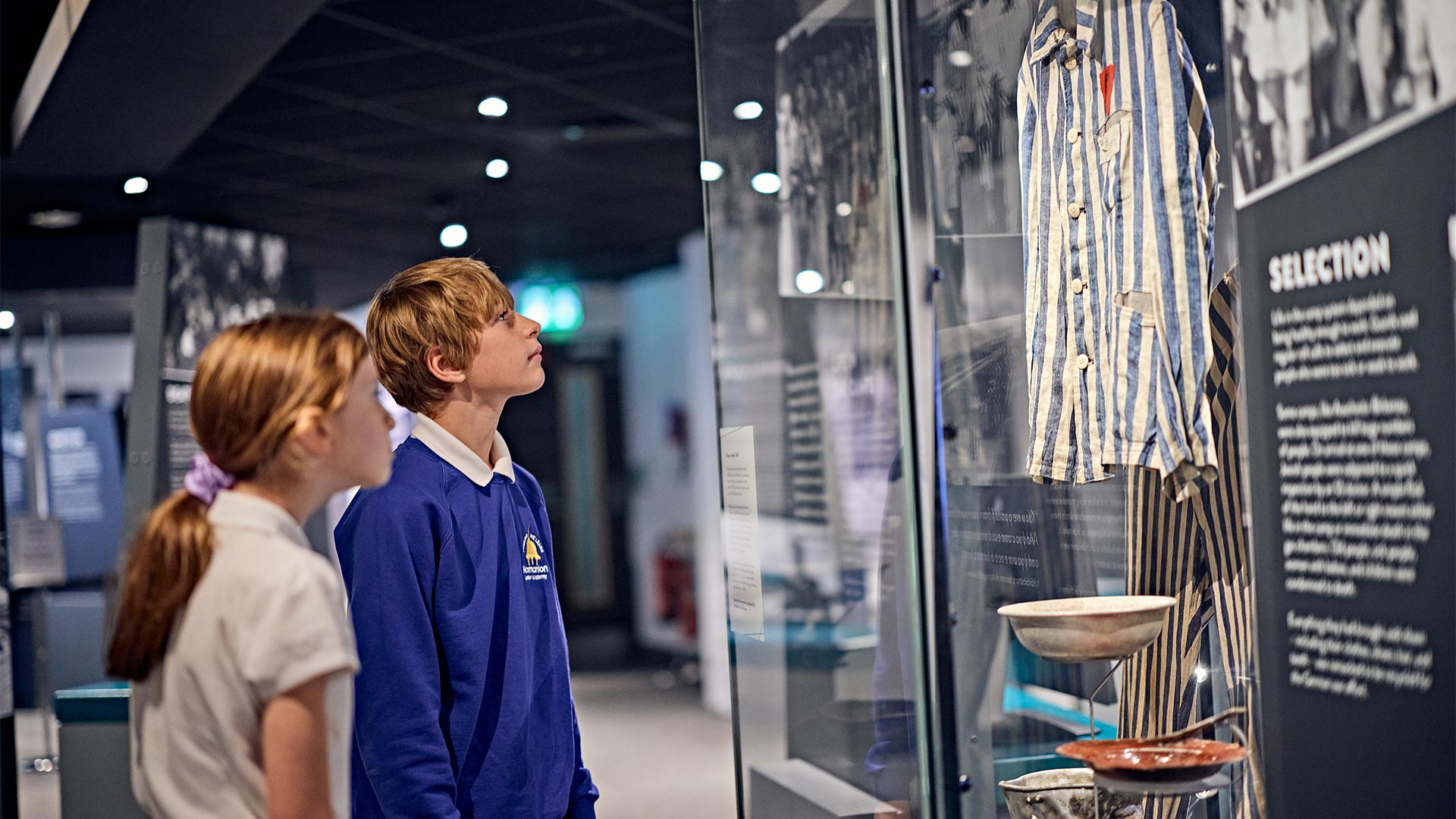Two schoolchildren stand in a museum gallery, observing striped clothing displayed in glass case, with historical artefacts and interpretive panels visible in a dimly lit exhibition space.